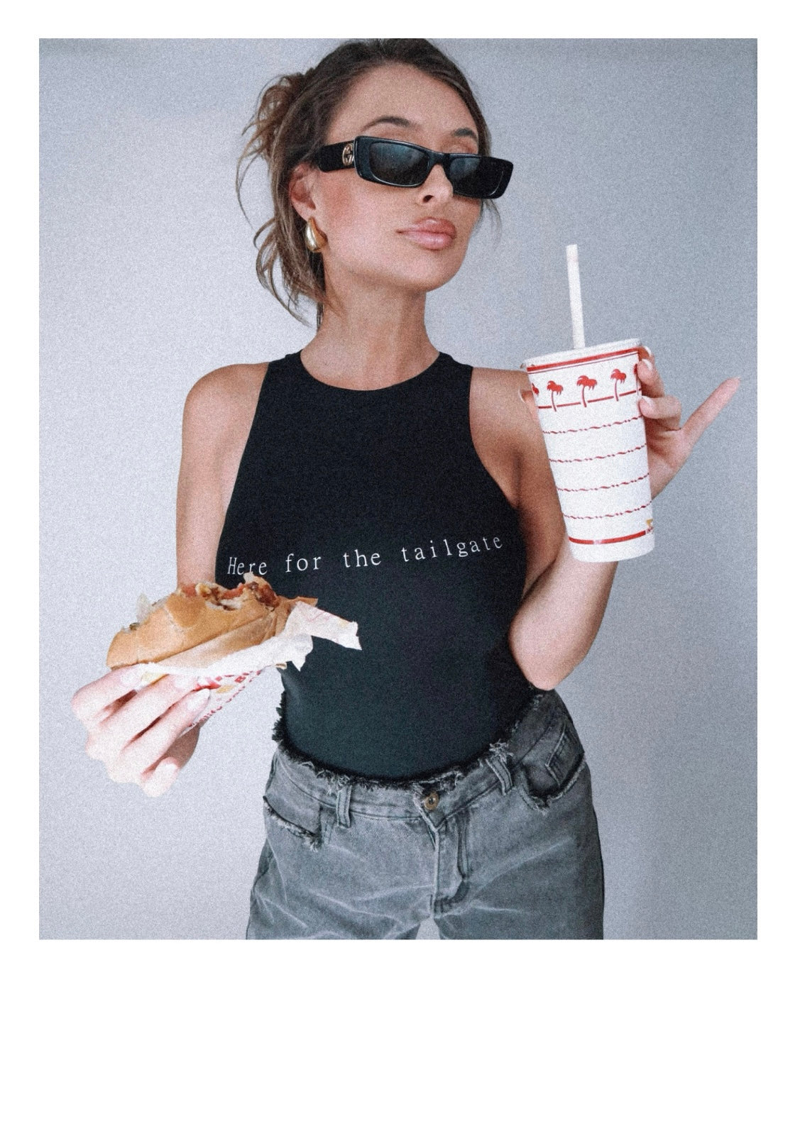 Model Eileen Hayman wearing Here for the tailgate high neck bodysuit in black, showcasing a sleek-form fitting design with flattering high neckline. Paired with distressed denim for a sporty, chic look, perfect for game day. Eating In-N-Out burger